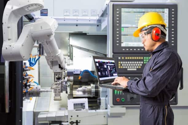 Engineer using a laptop to control a CNC lathe with robotic part handling for automated production