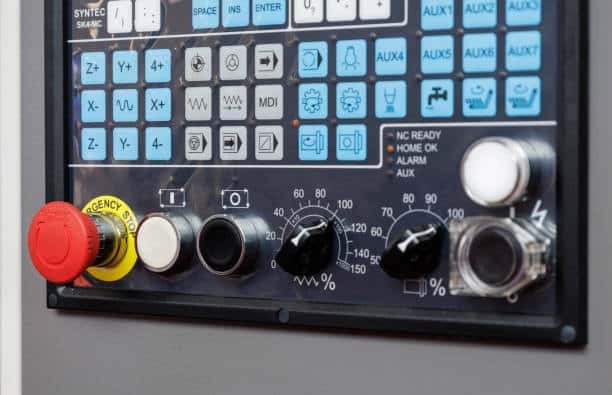 Image of a CNC control panel with a digital display, various buttons labeled Z, Y, X, and auxiliary functions, plus an emergency stop button, speed adjustment dials, and status indicators.