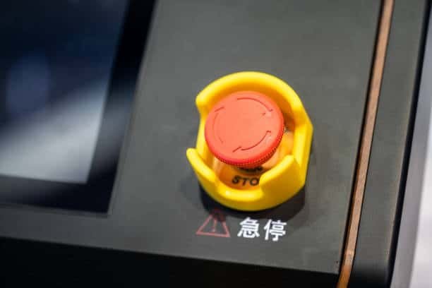 Close-up image of a red emergency stop button encased in a yellow guard on a black CNC machine panel, with a digital display in the background.
