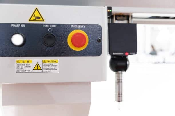 Image showing a red emergency stop button, power on/off switches, and a caution label on a white CNC machine control panel, with a cutting tool visible above.