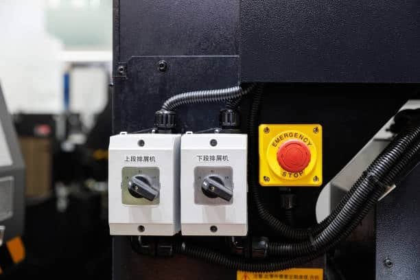 Image showing a red emergency stop button in a yellow casing next to two power switches on the side of a black CNC machine, with cables and industrial equipment in the background.