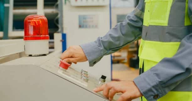 Photo of a worker wearing a high-visibility vest pressing a red emergency stop button on a CNC machine, with a red alarm light visible in the background.