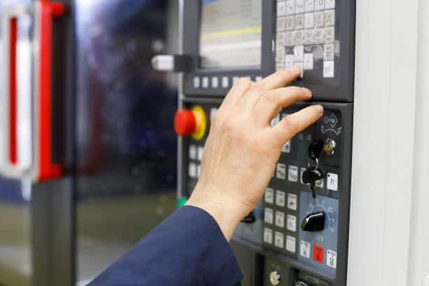 Image of a hand in a blue sleeve adjusting a knob on a CNC machine control panel featuring a digital screen, various buttons, and a red emergency stop button.