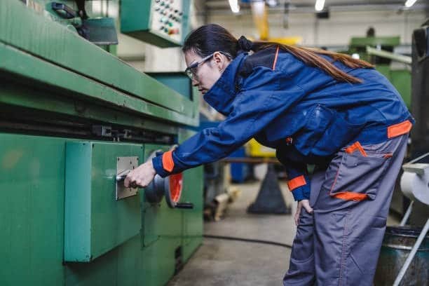 Image of a worker wearing a blue uniform with orange accents adjusting a lever on a green CNC machine, with industrial equipment visible in the background.