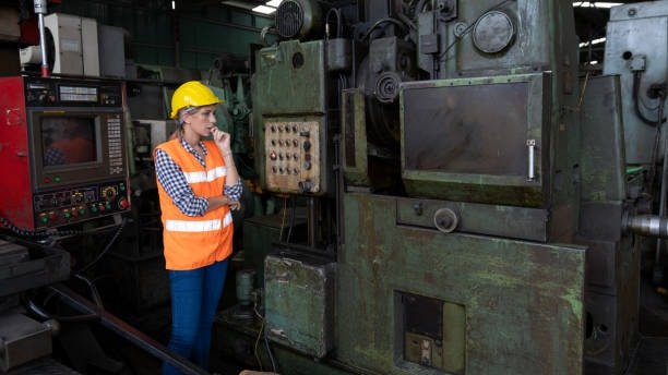 Female operator in high-visibility vest inspecting control panel near old CNC machine to assess vibration or chatter
