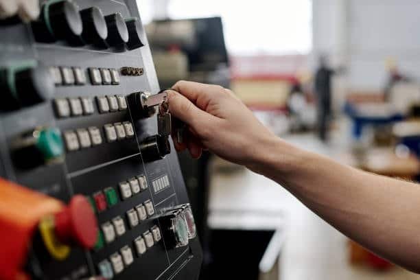 Photo of a hand turning a key in a lock on a CNC machine control panel with multiple buttons and switches, set against a blurred industrial background.