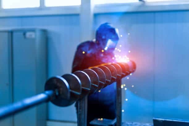 Automated welding process on a metal auger shaft with sparks and blue welding arc visible
