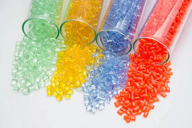 ABS plastic granules in green, yellow, purple, red, and blue spilling from glass tubes in a lab setting.
