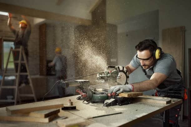 Construction worker cutting material with a miter saw, creating heavy dust in workshop