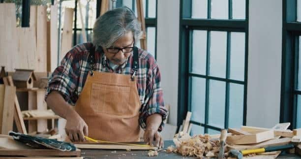Elderly craftsman measuring wooden board with tape before cutting in workshop