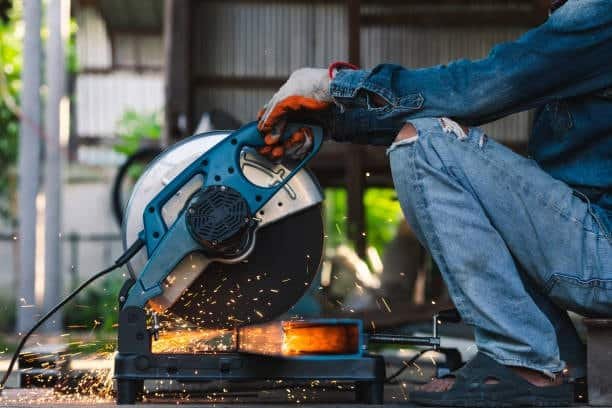 Worker using circular saw to cut metal pipe outdoors, with sparks flying during the cut