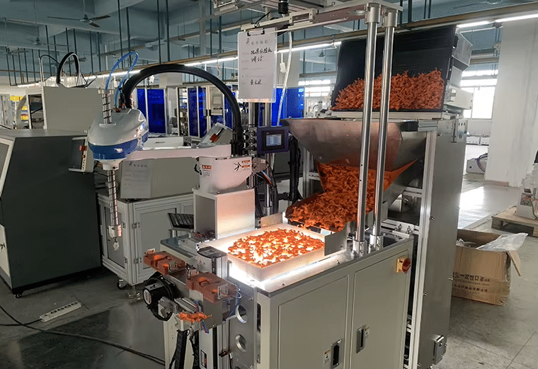 Automated gate trimming station removing excess material from orange injection molded plastic parts using a robotic arm in a factory setting.
