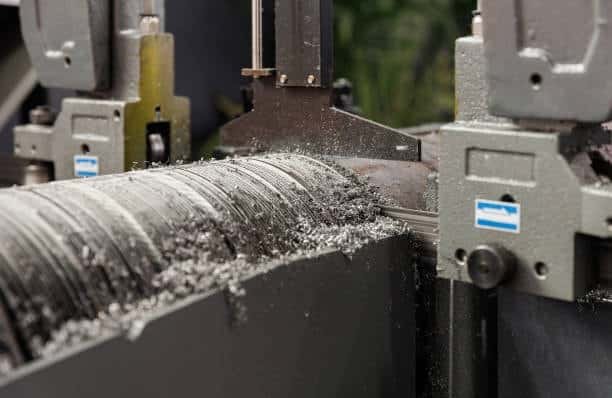Close-up of a horizontal band saw cutting a thick cylindrical metal rod with shavings