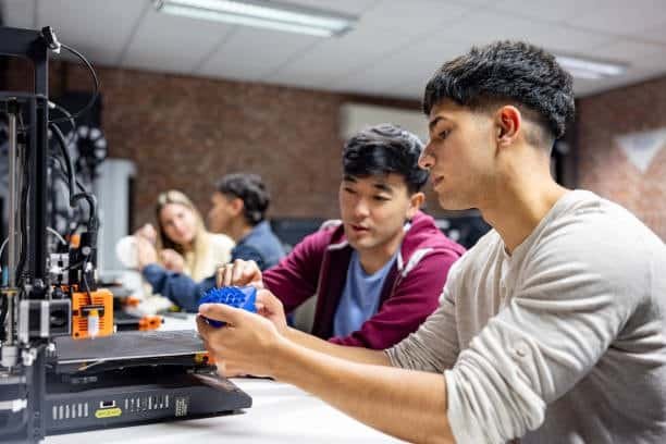 Students examining a 3D printed model fresh from an FDM printer in a classroom.Educational environments use FDM 3D printers to teach CAD modeling and material science.