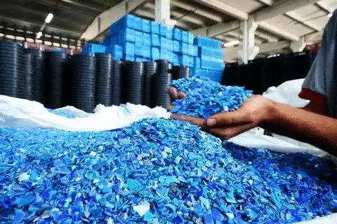 Worker handling a pile of blue recycled plastic flakes in a factory, showing raw material for remanufacturing.